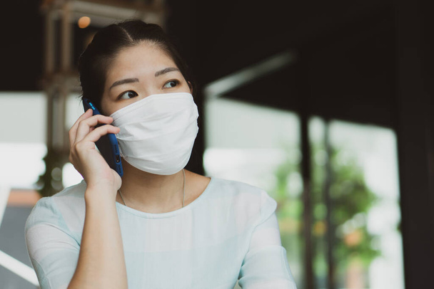 young asian woman wearing face mask and headphone and using computer to work from home during covid-19 or coronavirus outbreak. social distancing and new normal lifestyle comcept - Photo, Image