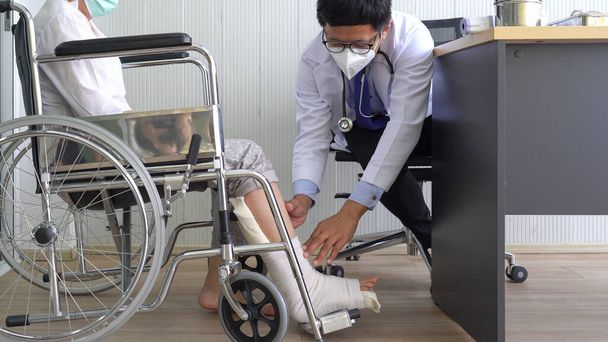 Asijský lékař v masce vyšetřuje pacientku, která sedí na invalidním vozíku, protože má poraněnou nohu. zdravotní péče a lékařské koncepce - Fotografie, Obrázek