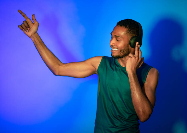 Αφρικανός αθλητής σε ακουστικά που χορεύει απολαμβάνοντας τη μουσική πάνω από το μπλε φόντο - Φωτογραφία, εικόνα