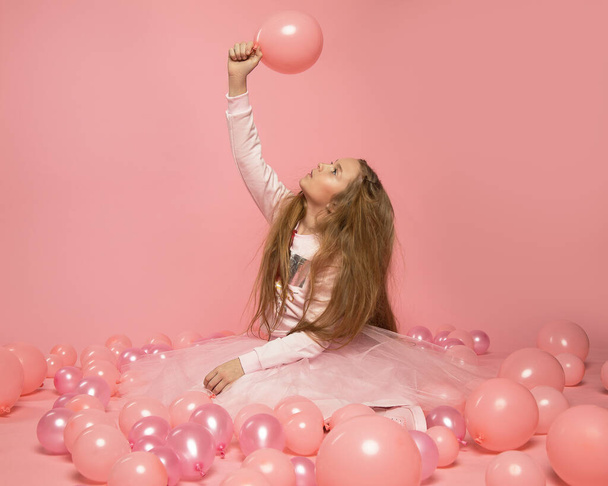 Beautiful funny 10-year-old girl with long fluffy hair in fashionable clothes is sitting on pink background in the Studio among inflatable balloons. The concept of the celebration. Teen glamour photo - Φωτογραφία, εικόνα