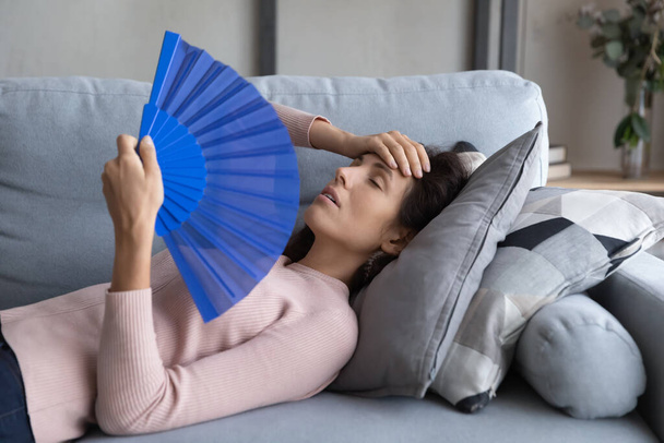 Overheated young woman relax on sofa wave with hand fan - Foto, imagen