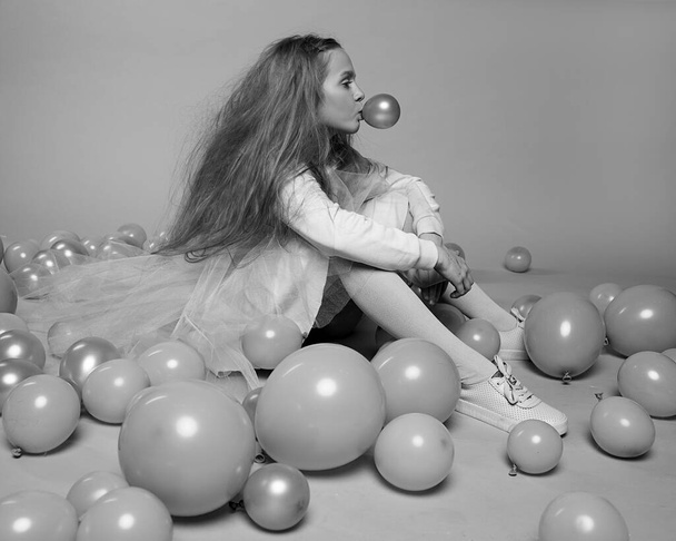A beautiful 10-year-old girl with long fluffy hair in fashionable clothes sits in Studio among balloons and inflates a bubble of chewing gum. The concept of the celebration. Black and white photo - Photo, Image