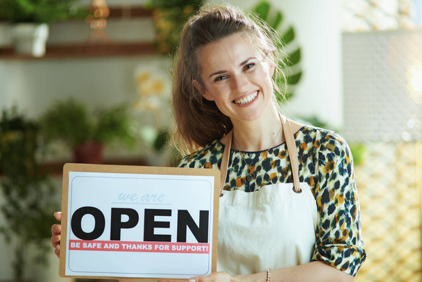 Opening small business after covid-19 pandemic. Portrait of happy modern 40 years old small business owner woman in apron showing open sign. - Фото, зображення