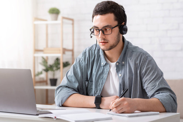 Leraar online. Man in bril en headset maakt notities in notebook en kijkt naar laptop - Foto, afbeelding