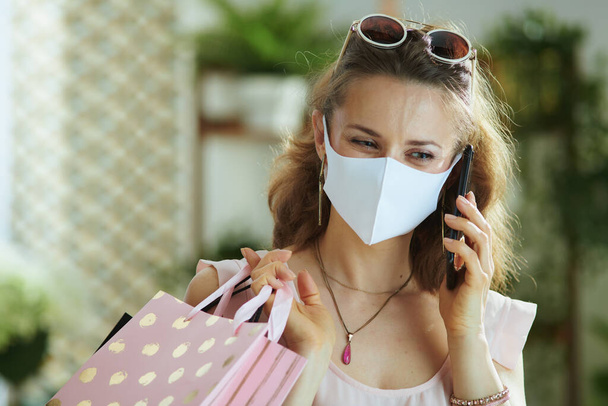 Life during covid-19 pandemic. trendy woman shopper in pink blouse with medical mask and paper shopping bags speaking on a smartphone. - Foto, Imagem