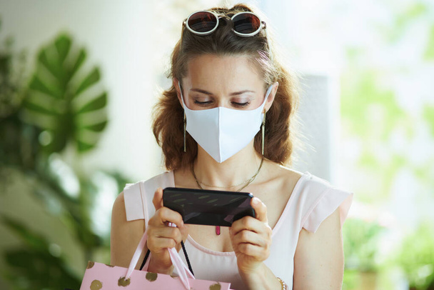 Life during coronavirus pandemic. stylish 40 years old woman shopper in pink blouse with medical mask, paper shopping bags and smartphone making online purchases on e-commerce website. - Valokuva, kuva