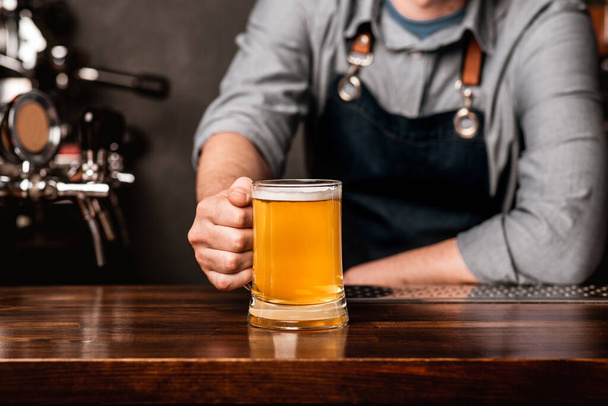 Freundlicher Barkeeper. Mann in Schürze stellt einen Becher Bier auf Holztheke im Innenraum - Foto, Bild