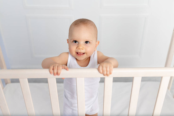 ritratto di un bambino sorridente di 8 mesi in piedi in una culla in camera dei bambini in abiti bianchi e guardando la fotocamera, bambino del mattino, concetto di prodotti per bambini - Foto, immagini