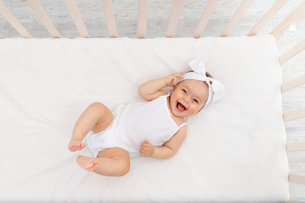 menina 6 meses de idade encontra-se em um berço no berçário com roupas brancas nas costas e ri, olha para a câmera, manhã do bebê, conceito de produtos para bebês
 - Foto, Imagem
