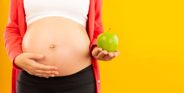 Happy pregnant young asian mother touch belly and holding fruit or green apple. Apple has vitamin, good for fetus, baby and pregnant woman with isolated on yellow background, copy space. Health care - Photo, Image