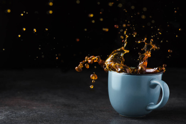 Studená káva stříkající v modrém šálku na kamenném stole, černé pozadí. Koncept čerstvého nápoje - Fotografie, Obrázek