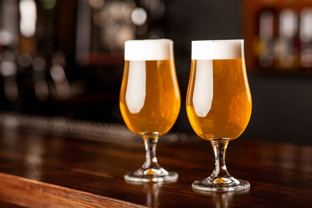 Ελαφριά μπύρα για πελάτες. Δύο ποτήρια σε ξύλινο πάγκο μπαρ στο θολό εσωτερικό - Φωτογραφία, εικόνα