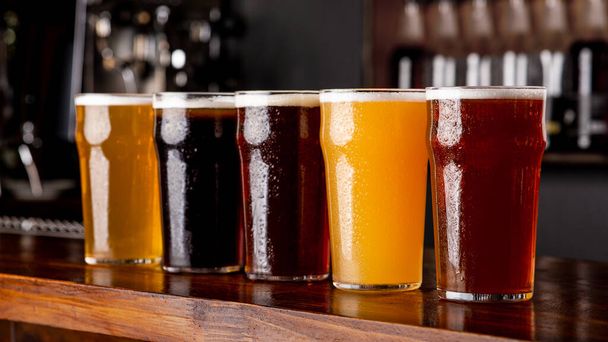 Нефильтрованное, темное, светлое пиво и эль в больших стеклянных бокалах на деревянной стойке в интерьере паба
 - Фото, изображение