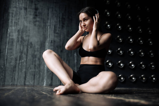 Yoga trainer is engaged in meditation in a sports uniform sitting in a lotus position and holding headphones with music in the gym. - Foto, afbeelding