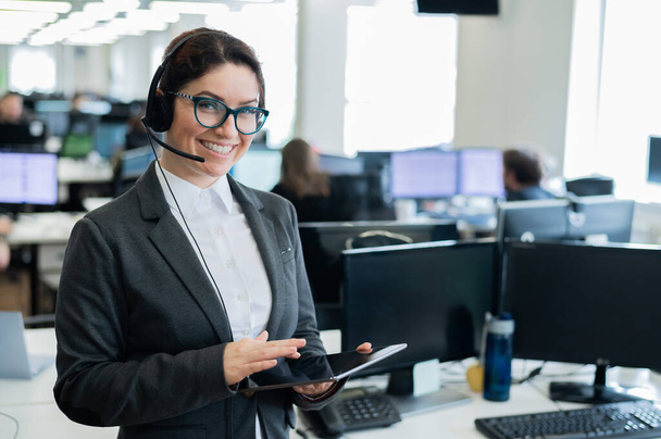 Beautiful caucasian woman in headset is holding a digital tablet while standing in open space office. A friendly female support service operator browsing device screen at the workplace. - Valokuva, kuva