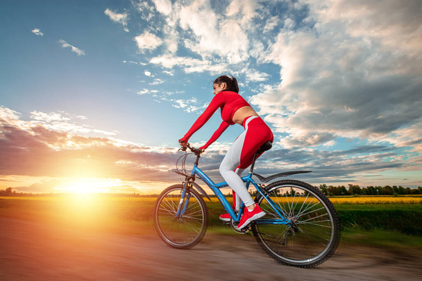 Beautiful girl in a red sports suit on a bicycle on a sunset background. The concept of a healthy lifestyle, sports training, cardio load. Copy space - Фото, изображение