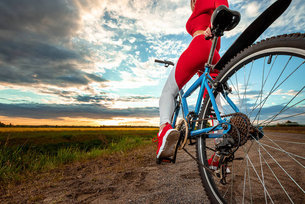 Beautiful girl in a red sports suit on a bicycle on a sunset background. The concept of a healthy lifestyle, sports training, cardio load. Copy space - Foto, immagini