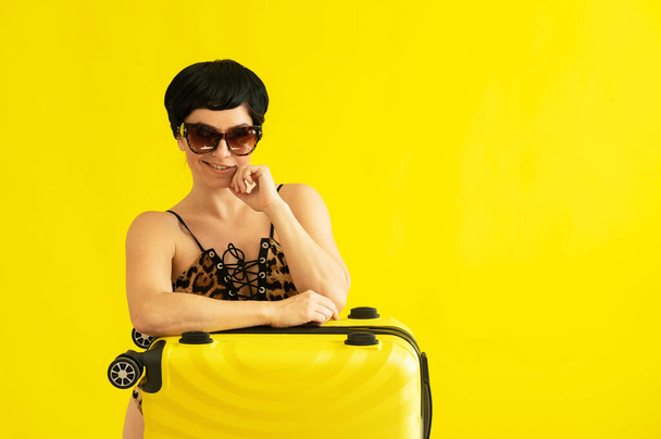 Portrait of an attractive woman in a one-piece leopard swimsuit and sunglasses with a suitcase on a yellow background. Seductive brunette in a monokini is ready to travel to the sea. Summer vacation. - Foto, Bild