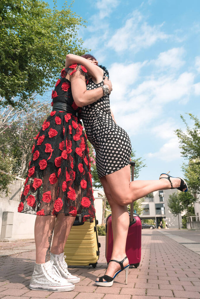 Dwie seksowne kobiety z walizkami obejmującymi się na kwiecistej ulicy miasta. - Zdjęcie, obraz