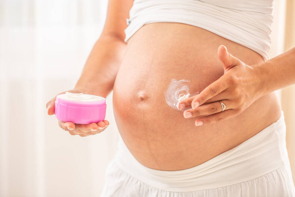Pregnant woman's belly detail, one hand holding a moisturizer and the second applying the lotion on the skin. - Fotografie, Obrázek