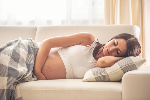 Kobieta w ciąży śpiąca na kanapie przykryta kocem, trzymająca jedną rękę na brzuchu. - Zdjęcie, obraz