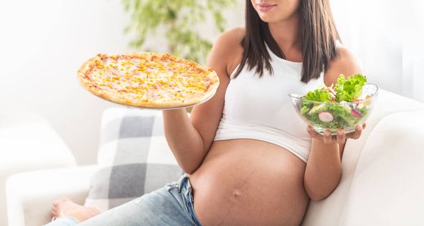 Zwanger meisje zit op een bank met salade kom in de ene hand en pizza in de andere. - Foto, afbeelding