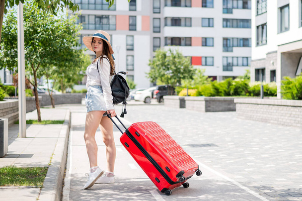 Beautiful young caucasian woman in a summer hat posing with a red suitcase outdoors. Happy smiling girl goes on vacation with a large luggage bag. Charming lady in shorts on a hot sunny day. - 写真・画像