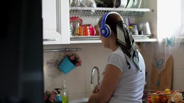 young woman washes the dishes listening to music with headphones - Video, Çekim