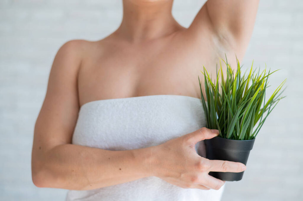 Faceless woman in a white towel holds a pot with a plant. Unrecognizable girl with the help of green bushes imitates the vegetation of armpit hair. Depilation area. Female hair removal. - Photo, Image