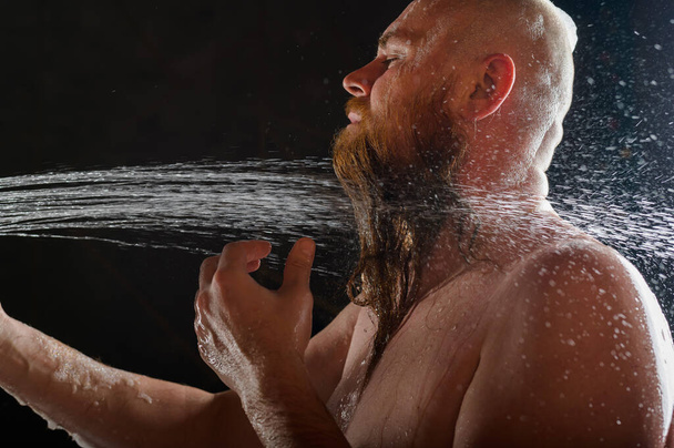 Чоловік приймає душ. Жорстокий чоловік з червоною бородою стоїть у ванній кімнаті під водопроводом і миє. Розсіювання спрею на чорному тлі.. - Фото, зображення