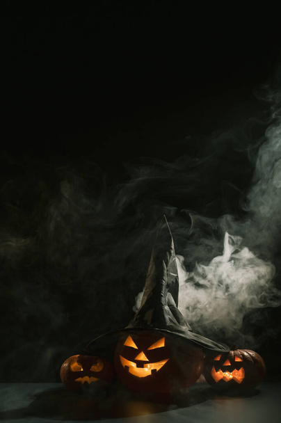 Vertical Halloween card. Witch hat on a pumpkin with carved creepy grimaces on a black background in the fog. Jack-o-lantern glows from the inside and emits smoke. - 写真・画像