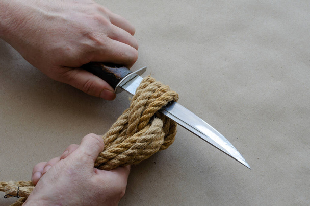 Handen van een volwassen man beslissend gesneden met een mes een grote knoop van hennep touw. Man van Kaukasische afkomst, 44-45 jaar. Bovenaanzicht onder een hoek. Selectieve focus. - Foto, afbeelding