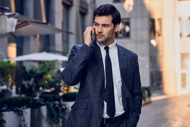 sebevědomý mladý muž v plném obleku mluví na mobilním telefonu a dívá se stranou, zatímco stojí venku s městskou krajinou v pozadí - Fotografie, Obrázek