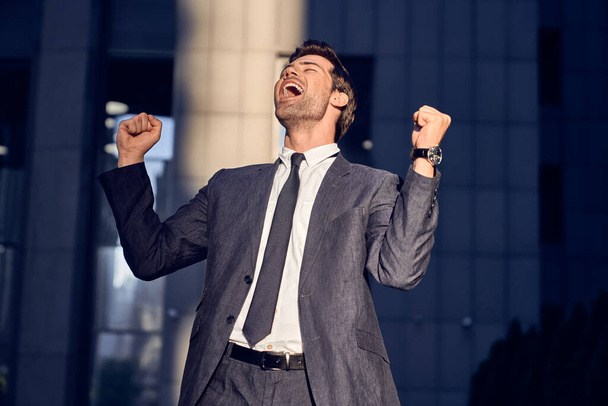 Yes, I did it. Happy young businessman keeping arms raised and expressing positivity while standing outdoors. - Foto, Imagen