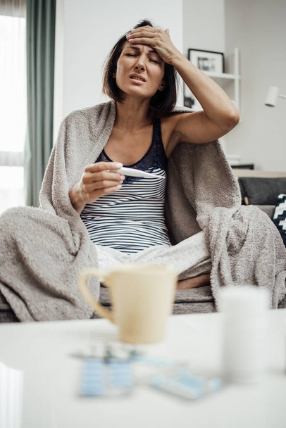 Sick and exhausted middle age woman suffering from flu fever. She is sitting on sofa at home, holding thermometer and measuring body temperature wrapped in beige blanket. - Foto, afbeelding