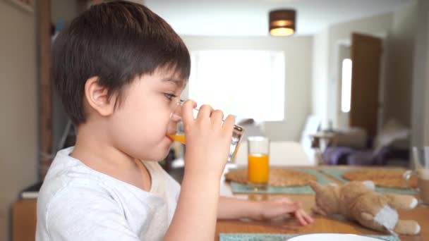 健康的な子供は透明なガラスからの新鮮なオレンジジュースを飲み、彼のおもちゃと遊ぶ。家で魚の食糧と朝食を持っている子供の男の子との新しい正常な健康な生活。食卓での家族の時間 - 映像、動画