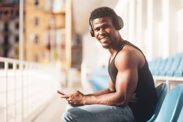 アフリカ系アメリカ人の男性が夏に屋外でヘッドフォンで音楽を聴いたり笑ったりスマートフォンやヘッドフォンでスポーティ男をコピーしたり - 写真・画像
