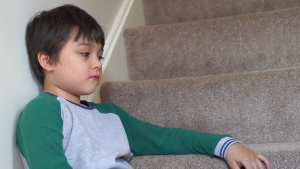 Gyerek fiú szomorú arccal ül a lépcsőn, szomorú gyermek néz le a gondolkodó arc, mentális egészség gyermek - Felvétel, videó