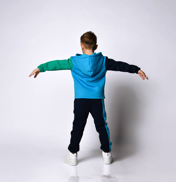 Rear view of little boy model in fashionable cotton sportsuit doing exercises spreading arms and legs. Full length portrait isolated on light grey - Foto, afbeelding