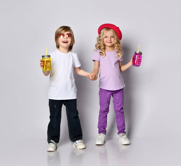 Little girl and boy in colorful casual clothes. Smiling, holding hands, yellow and pink cocktail bottles. Posing isolated on white - 写真・画像