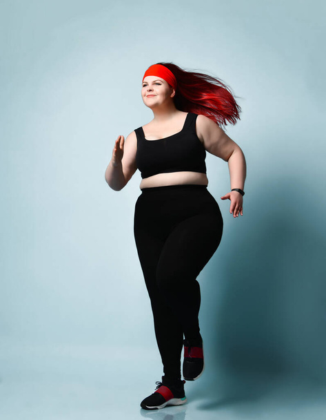 Happy smiling red-haired plump lady running in place on blue background. Fitness, physical activity, body positive. Full length portrait, copy space - Фото, зображення