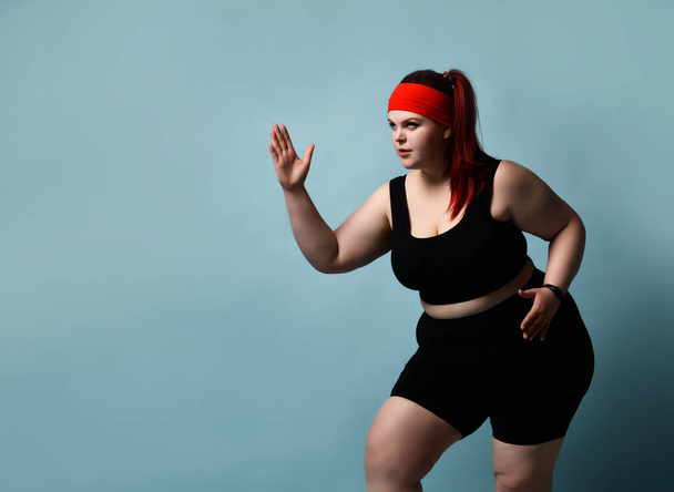 Serious concentrated young plus-size lady does exercises looking up with legs flexed and arms bent in elbows on blue background. Fitness. Copy space - Foto, Bild