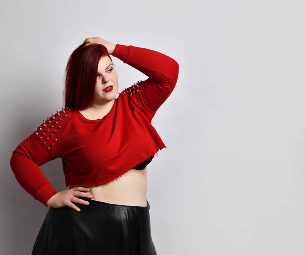 Overweight redhead woman in red spiked top, black leather skirt. Touching face, put hand on hip, posing sideways isolated on white - Photo, image
