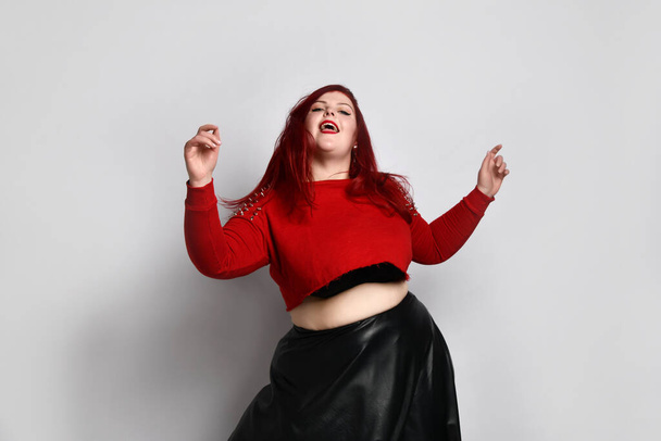 Obese ginger lady in red spiked top, black bra and leather skirt. She is dancing, posing isolated on white photo background - Foto, Bild