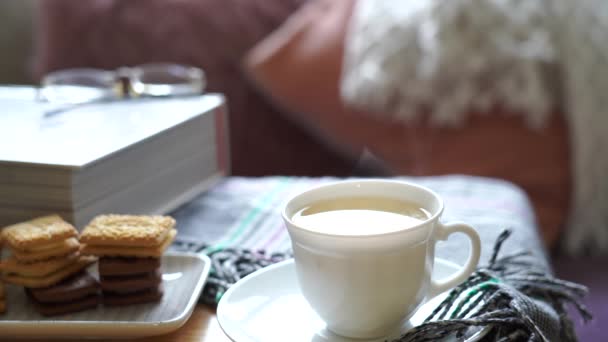 Forró csésze tea gőz az asztalon homályos párnák kanapé hátterén.Jelenet hangulatos és pihentető délutáni tea ropogós keksszel tálalva napsütéses napon tavasszal vagy nyáron, angol hagyományos  - Felvétel, videó