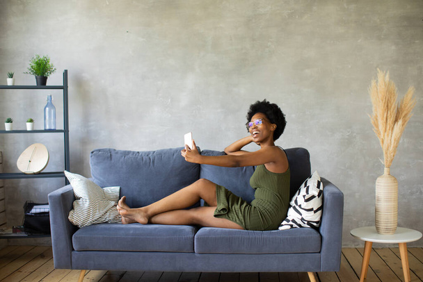 Happy african American young woman sit relax on cozy couch happy to move to new apartment. Smiling black girl rest on comfortable sofa in living room dreaming - Foto, imagen