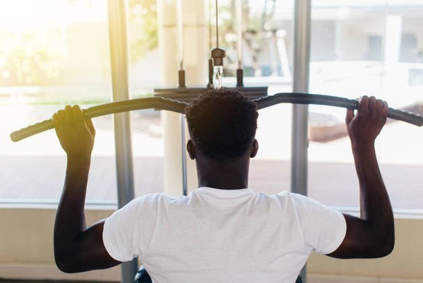 Strong African American man in sportswear pulling cable with bar while sitting on exercise machine during training in gym - Φωτογραφία, εικόνα