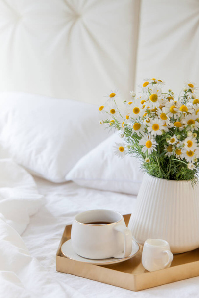 Morning breakfast in bed. White bedding. Golden tray in the nordic style. White cup with black coffee and a milkman with milk. Daisies flowers in a vase. White little alarm clock - Foto, Imagem