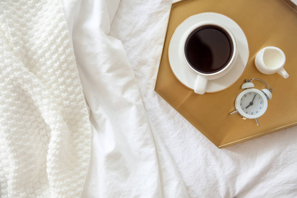 Morning breakfast in bed. White bedding. Golden tray in the nordic style. White cup with black coffee and a milkman with milk. Daisies flowers in a vase. White little alarm clock - Foto, imagen