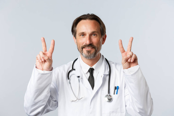 Covid-19, preventing virus, healthcare workers and vaccination concept. Cheerful handsome middle-aged doctor in white coat showing peace signs or v-sign and smiling, stay positive - Photo, Image